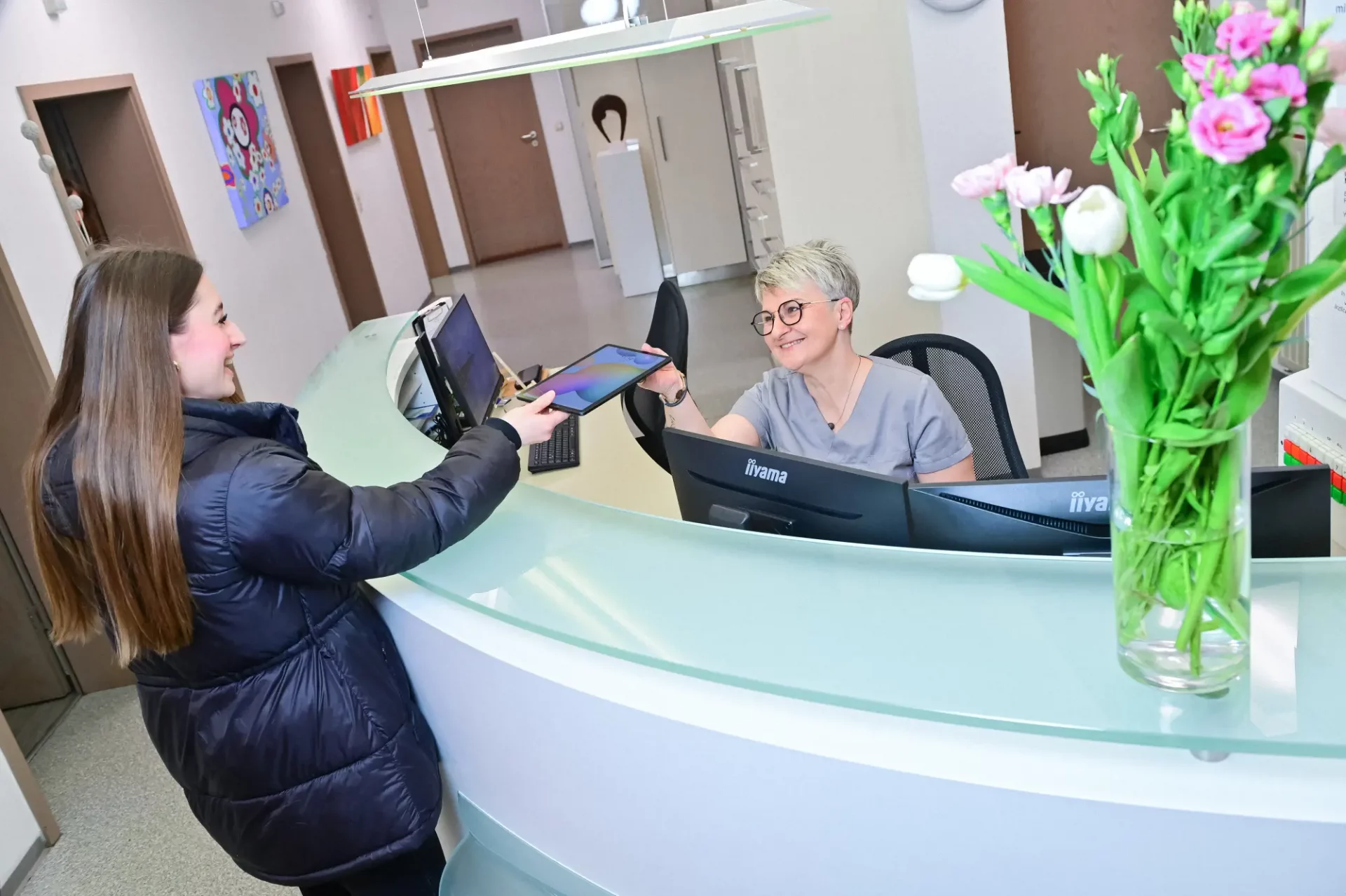 Eine Mitarbeitein am Empfangstresen überreicht einer Patientin ein Tablet.
